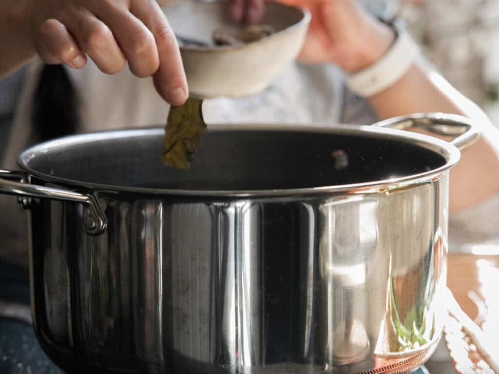 A person adds ingredients from a small bowl into a large stainless steel pot. The focus is on the hand holding a leaf above the pot. The scene suggests cooking or preparing a dish.